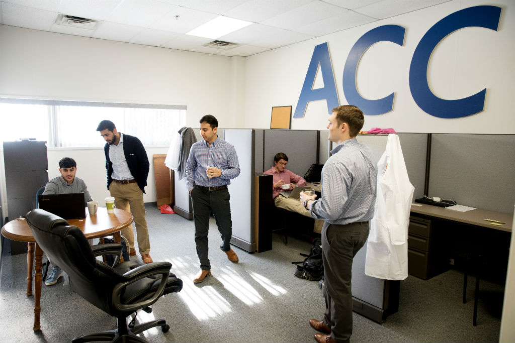 Sepehr Zomorodi, Ameer Shakeel, Payam Pourtaheri, Joseph T. Frank, and Zachery Davis begin their day discussing emails and work over coffee. On the wall above are the initials for the Atlantic Coast NCAA athletic conference, to which their alma mater, UVA, belongs. The team members are big supporters of UVA athletics.