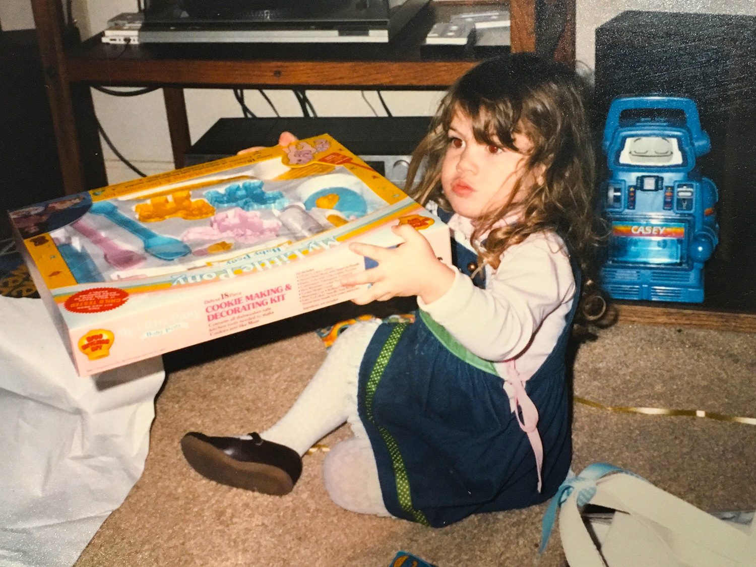 Sterling as a toddler sits on the ground holding a cookie making and decorating toy, still in packaging. Wrapping paper and ribbon are on the floor around her. 