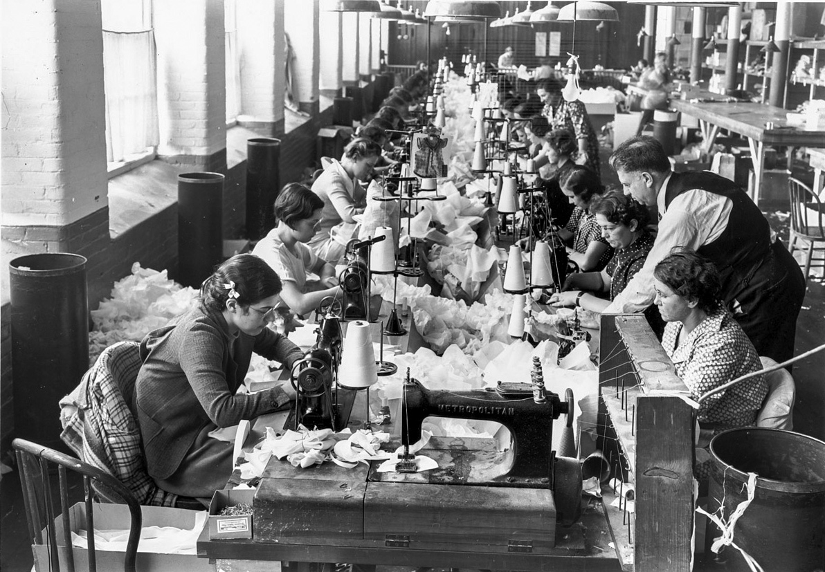 A long table with women sitting side-by-side working on sewing machines and surrounded by fabric 