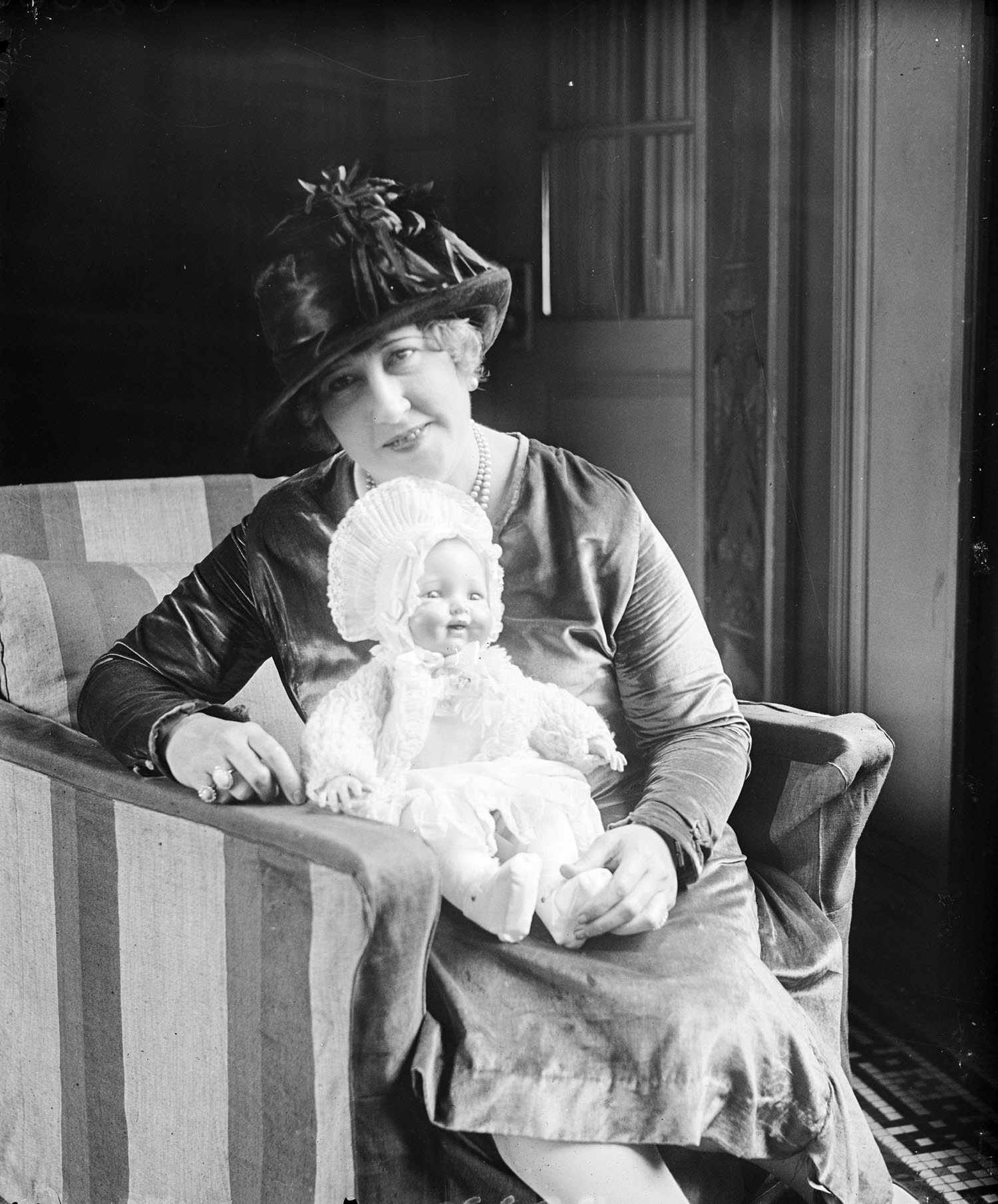 A woman, sitting in a chair smiling, wearing a fashionable hat and holding a lifelike baby doll 