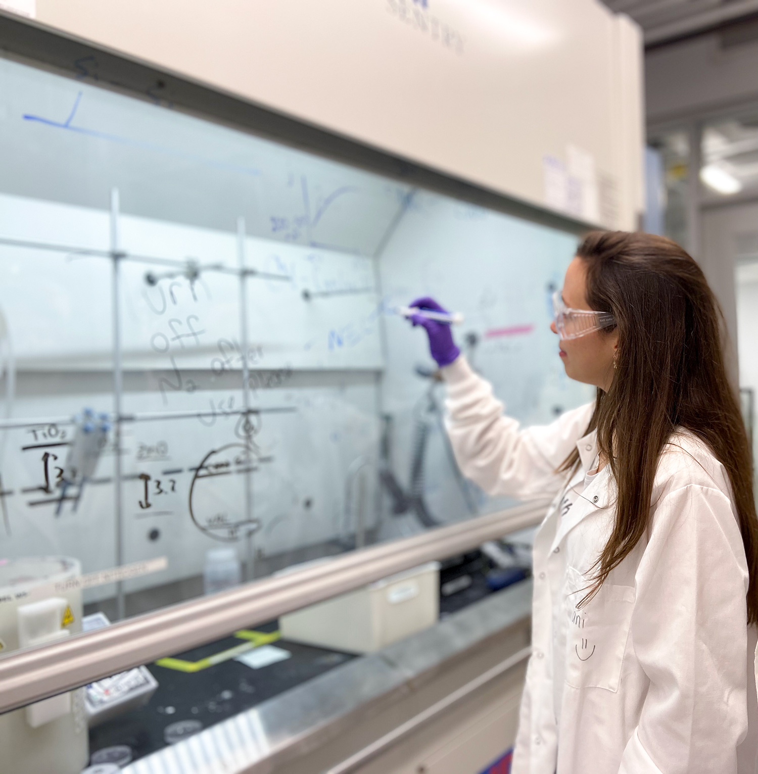 La ingeniera química Daniela Blanco escribe atentamente ecuaciones químicas en una ventana de vidrio dentro de un laboratorio de química. Lleva el atuendo tradicional de laboratorio: bata blanca, gafas de seguridad y guantes de plástico.