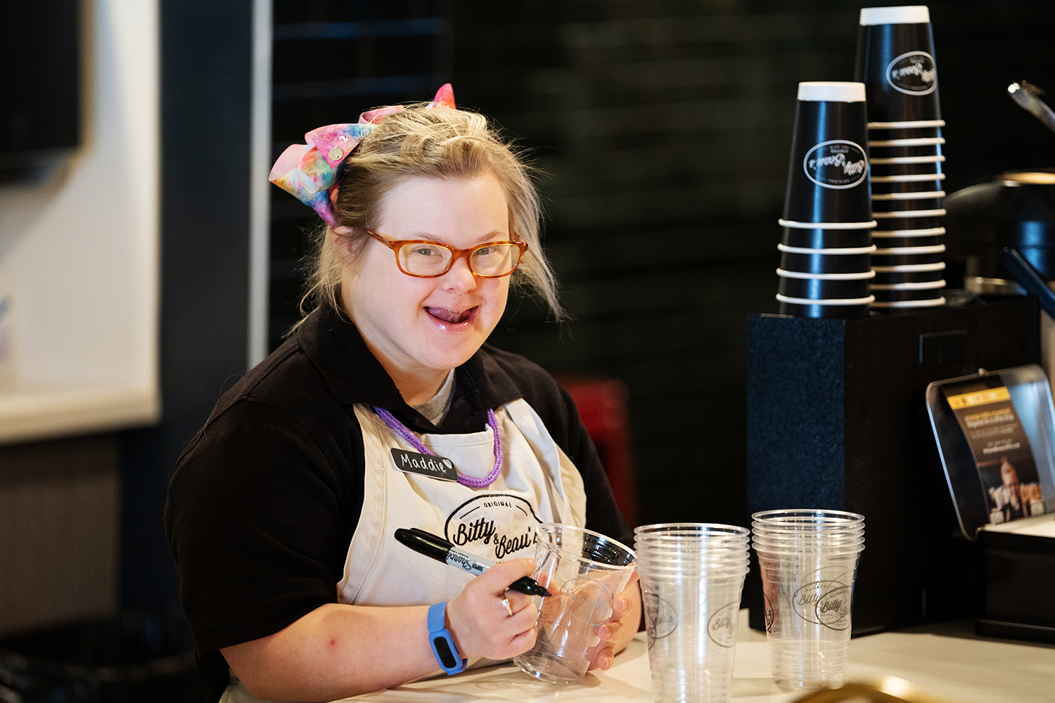 Female employee stands at a counter, smiling at the camera. She holds a plastic cup and a sharpie, with stacks of cups in front of her. She wears a black shirt, a beige apron with logo, and colorful accessories. 
