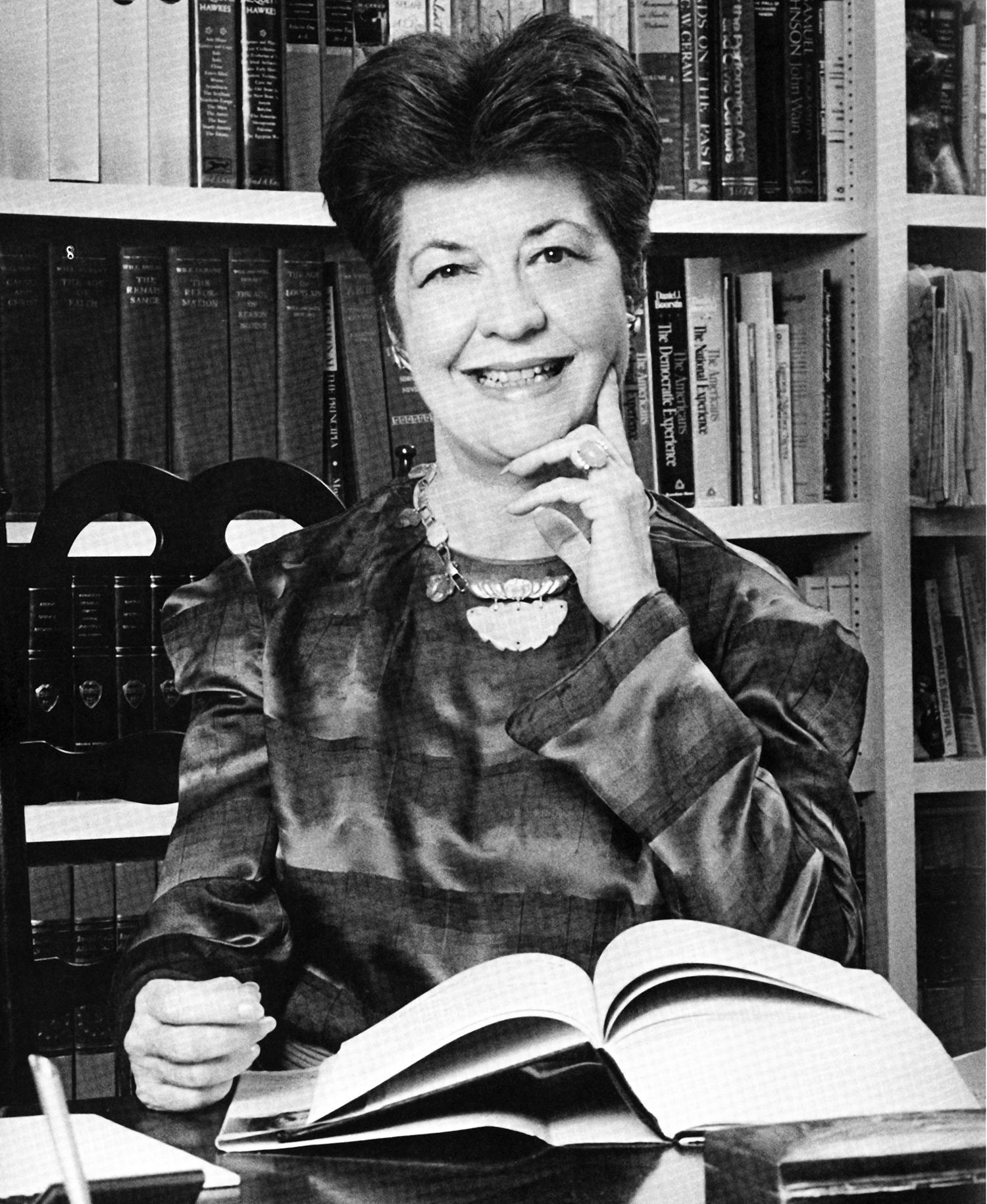 A black and white photograph of a smiling woman in business attire sitting at a desk with an open book. A bookshelf filled with books is seen in the background. 