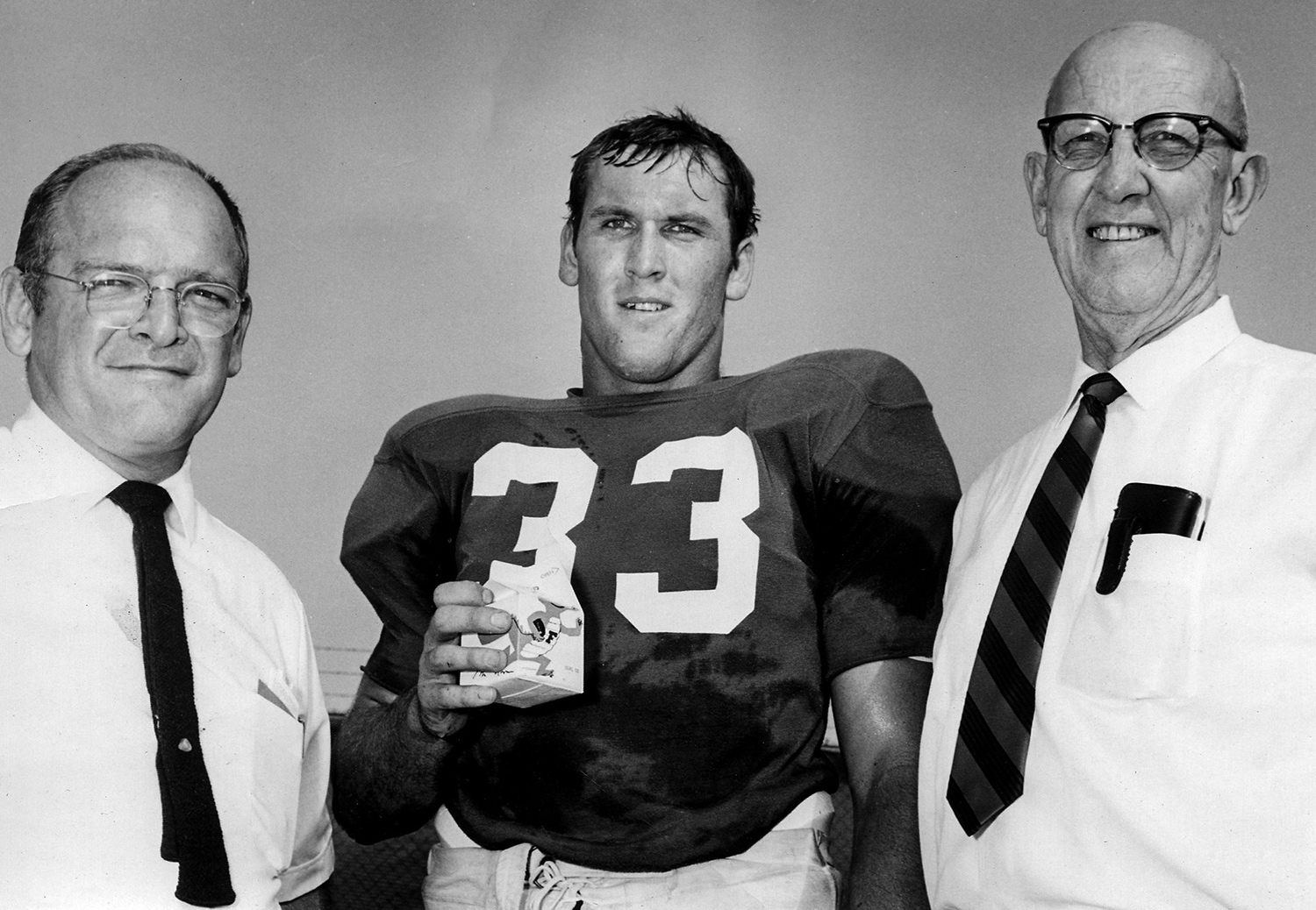 Football player holds carton of Gatorade and stands between two older men on a football field