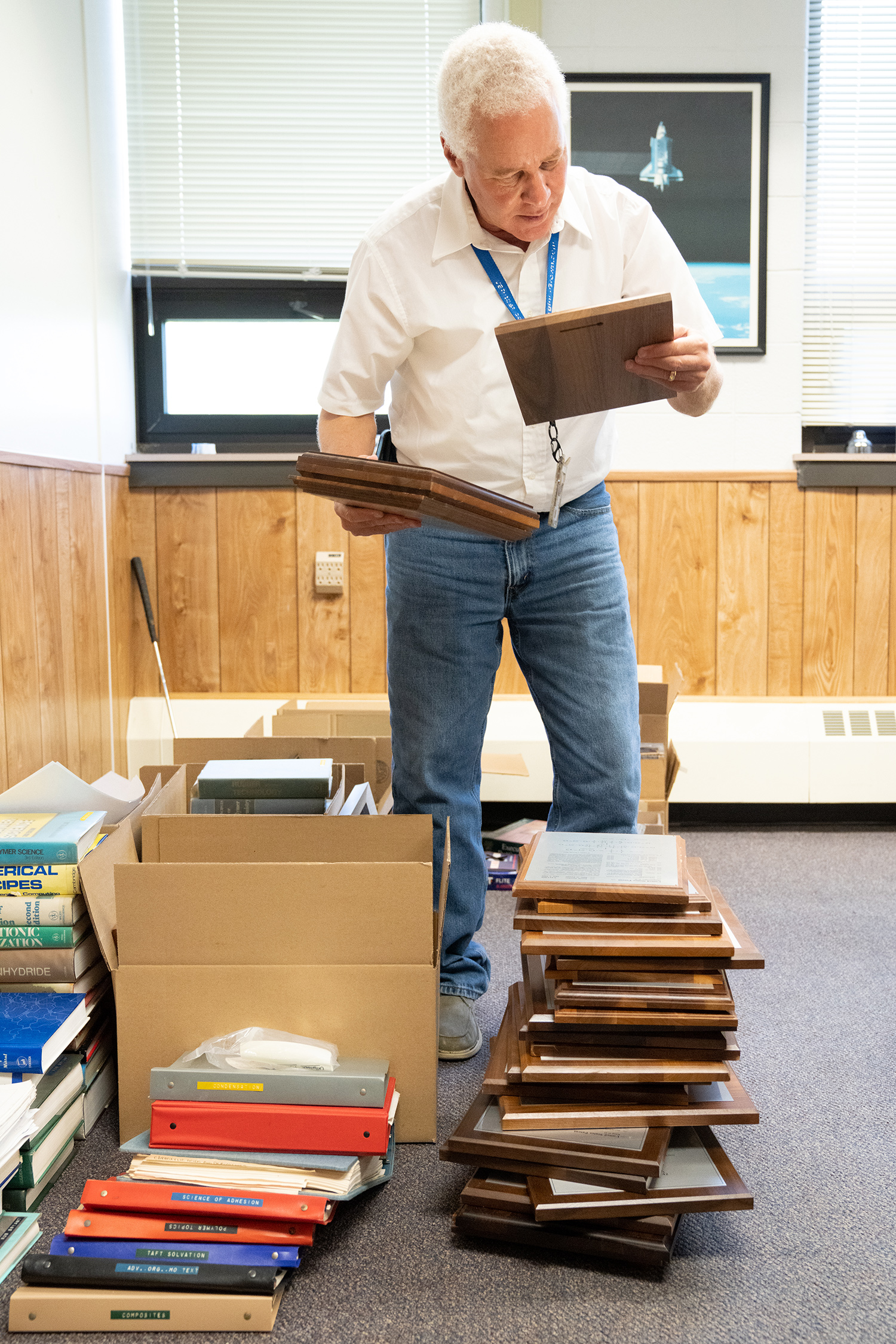 Dr. Robert Bryant smiling and in a white button up shirt and blue jeans with a blue lanyard around his neck looking at his plaques while surrounded by more plaques, binders, science books, and boxes in a wood-paneled office with dark multi-color carpet