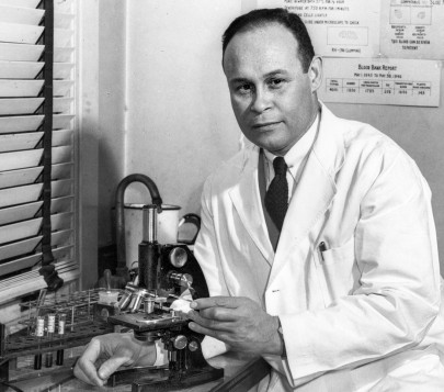 外科医生兼研究员查尔斯·德鲁博士身穿白色实验服，坐在显微镜前。