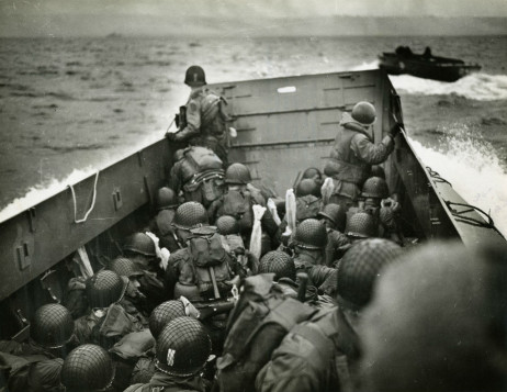 Image: Helmeted U.S. soldiers look out over the prow of a Higgens boat as it navigates crashing waves towards the beaches of Normandy on D-Day.