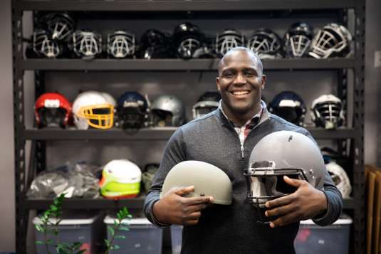 Shawn Springs holds two helmets and poses in front of a wall of helmets.