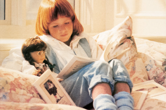 Bathed in sunshine, a young girl with a bowl-cut wearing a jeans jumper and socks, cozily curls up on a pillowy, pastel-floral window seat and reads to her doll from a book in her lap. Several additional books whose covers match the doll by her side are strewn about.