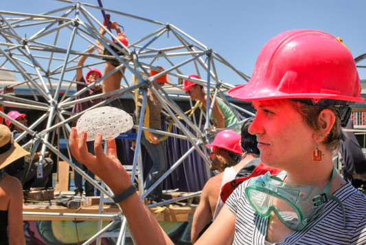 With a colorful purple buzzcut, tank top, and sunglasses, scientist Natalia Bilenko smiles in the sun and heat as she works alongside others to assemble metal struts and wiring to form the sculpture “Dr.Brainlove.” 