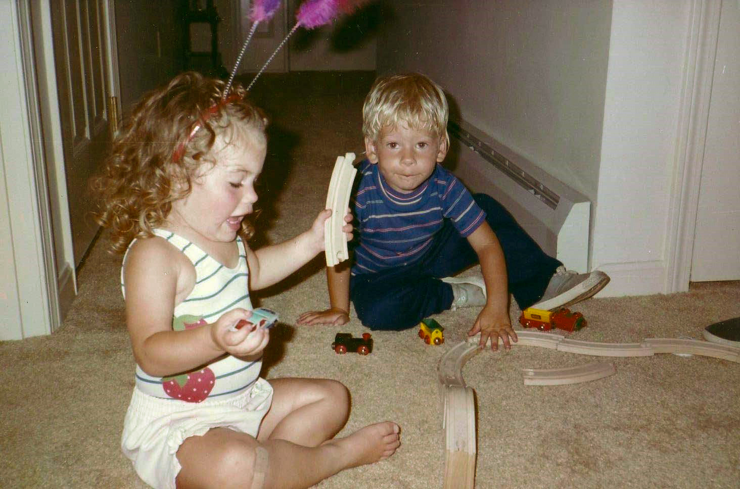Sterling as a toddler sits on the ground with a young boy. Toy cars and pieces of wooden track are around them on the floor and in Sterling's hands. An assembled track is in front of them. 