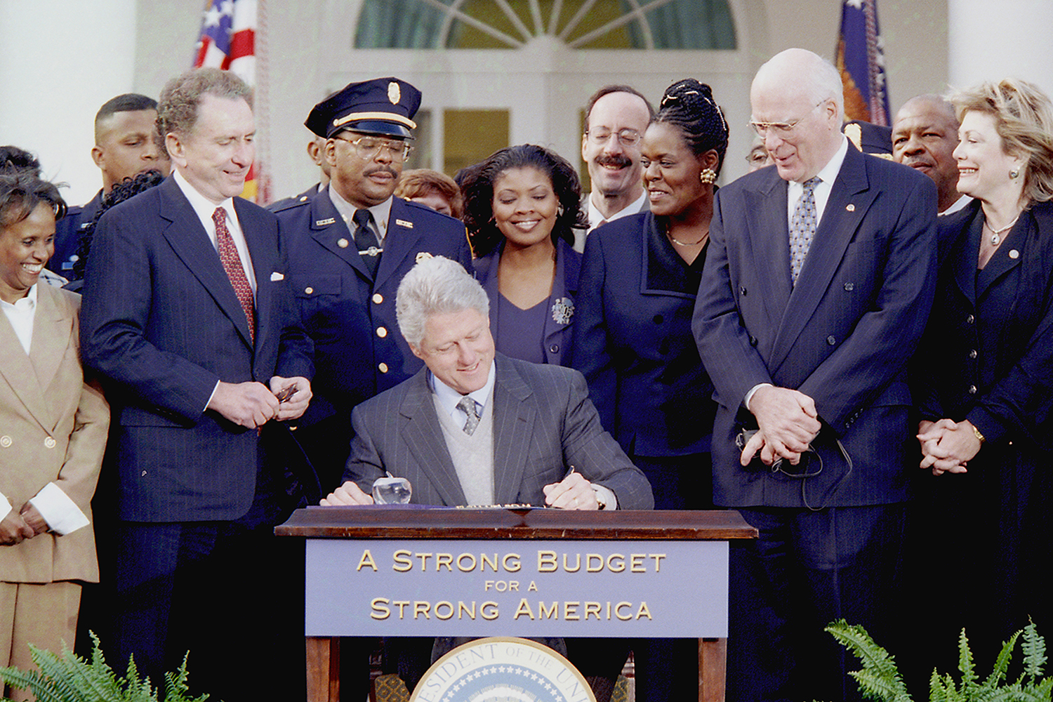 Then-President Bill Clinton signs an appropriations bill that includes the Patent and Trademark Office Efficiency Act into law, in the Rose Garden on November 29, 1999. Clinton is seated and pictured in the middle of the image, surrounded by police and other officials who are standing. The desk Clinton is using to sign the bill says on the front, 'a strong budget for a strong America.' The photo was taken by David Scull and provided courtesy of the William J. Clinton Presidential Library.