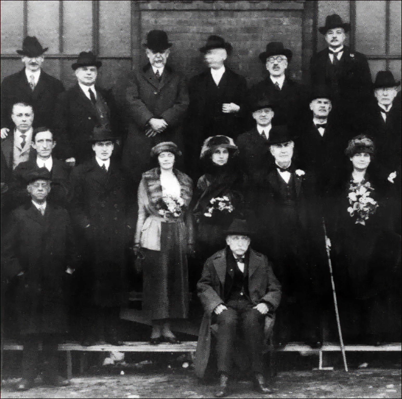 a group 18 well-dressed men and women are standing, and one man is sitting, in a photo taken in 1920. 