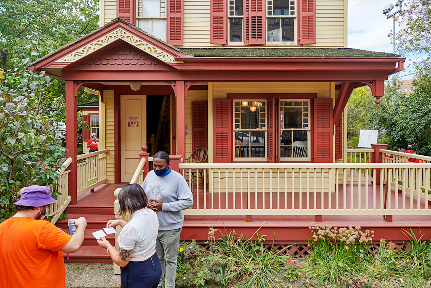 a tall man watches as a couple reads documents outside of a two-and-a-half story beige home with red trim. 