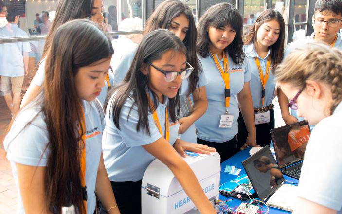 A number of high school students in light blue shirts interact with an individual at a display table. Before them is a white device, electronics and a number of laptops.