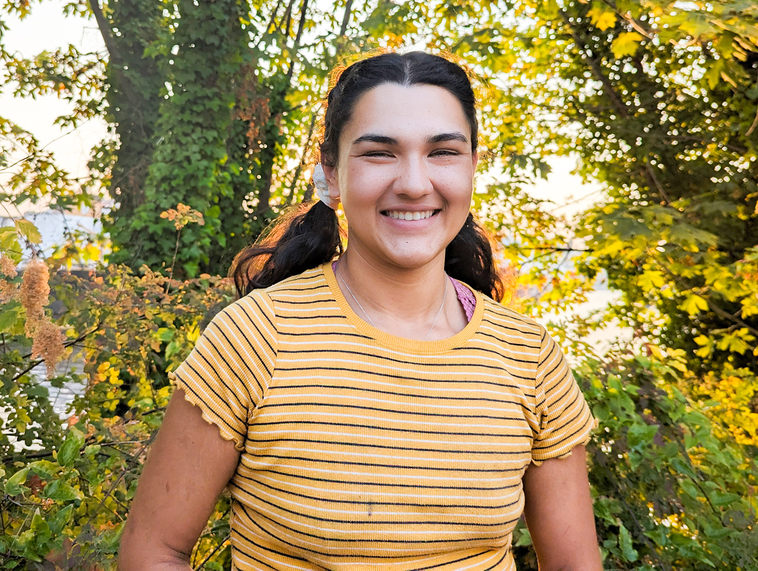 A woman wearing a horizontally striped yellow short sleeve shirt smiles at the camera with foliage in the background.