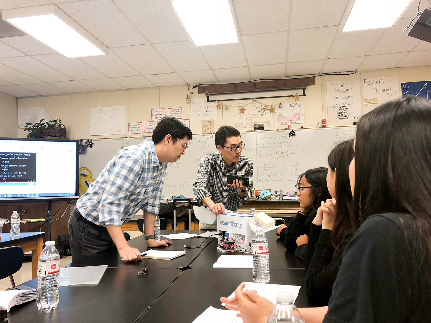 Two men talk to young high school females in classroom setting, while examining a device labelled Heart and Sole.