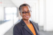 Sonia, a woman with dark eyes, glasses, and short gray-black hair, smiles in USPTO headquarters, wearing gold hoop earrings, a gold necklace, a peach shirt, and a black blazer with small white polka dots.