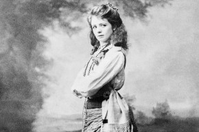 Maude Adams wearing a tunic, skirt, sash, and beads stands at three-quarters profile, framed by trees in the background, and looks off to the right.